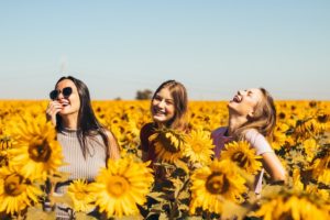 A imagem mostra três amigas rindo em um lindo campo de girassóis