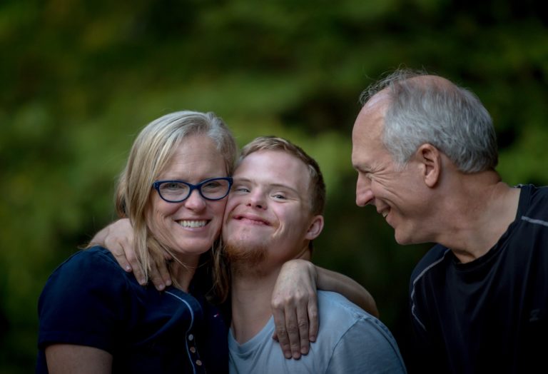 A imagem mostra uma pessoa com deficiência sorrindo ao lado de seus pais.