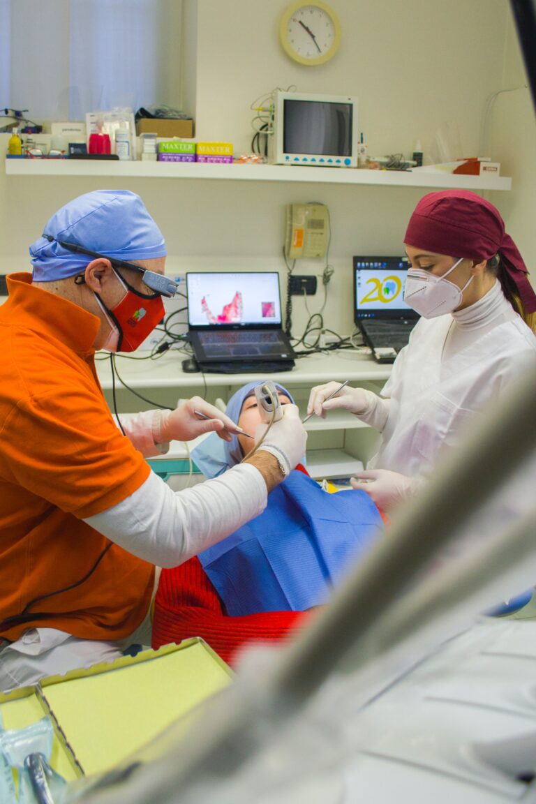 A imagem mostra um dentista e sua instrumentadora realizando procedimentos na boca de um paciente que se encontra deitado na maca