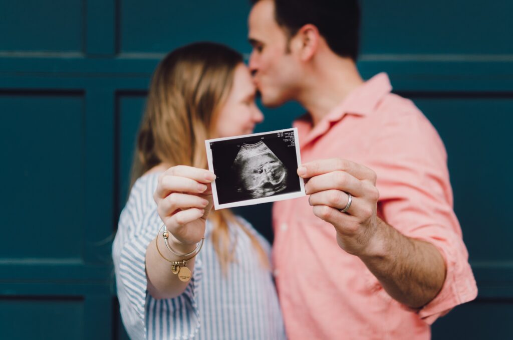 A imagem mostra um casal segurando uma foto de um ultrassom do bebê que eles esperam